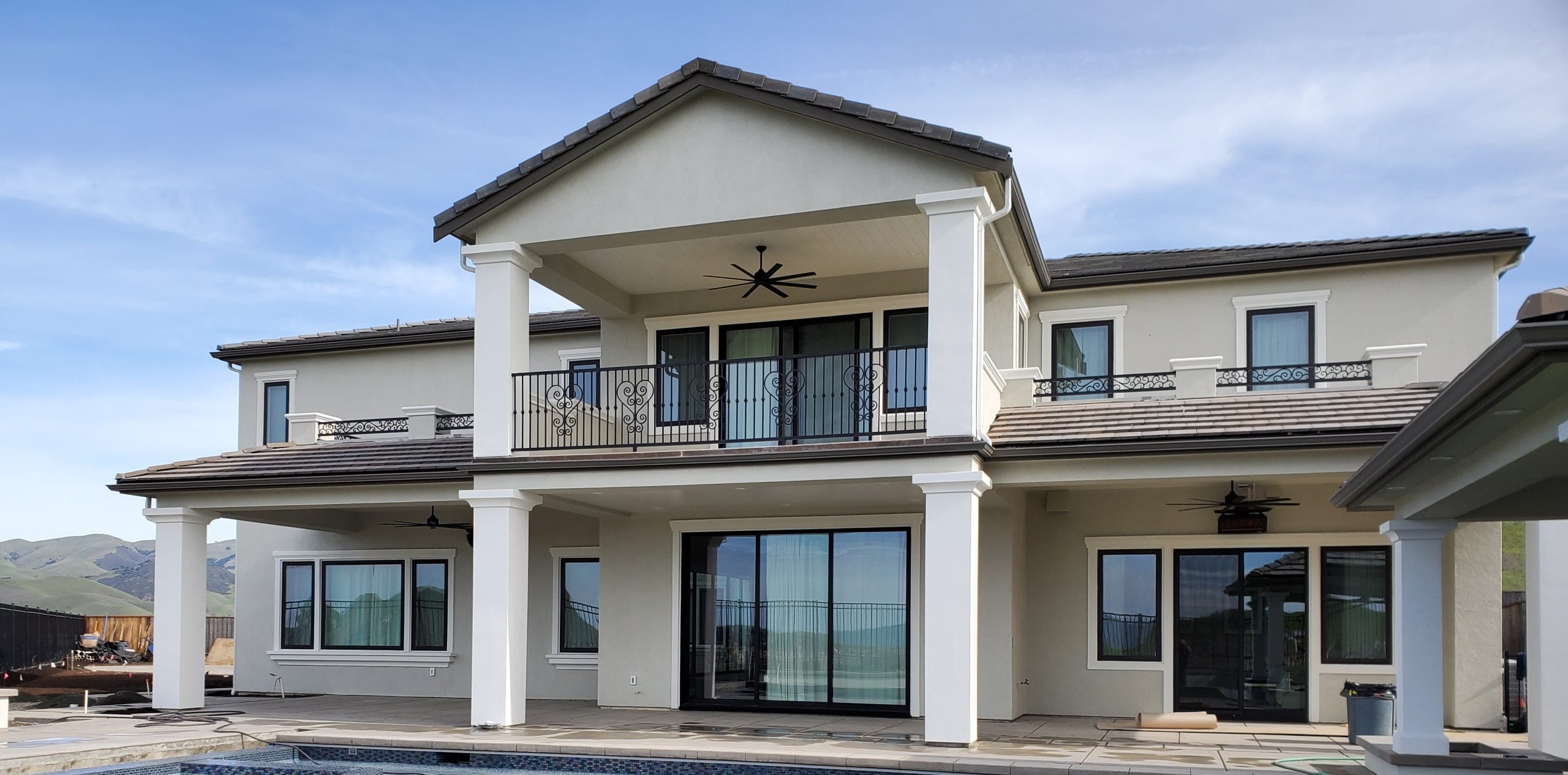 Backyard view of a modern two-story luxury home with white pillars, an upper balcony, and large glass doors next to a pool area.