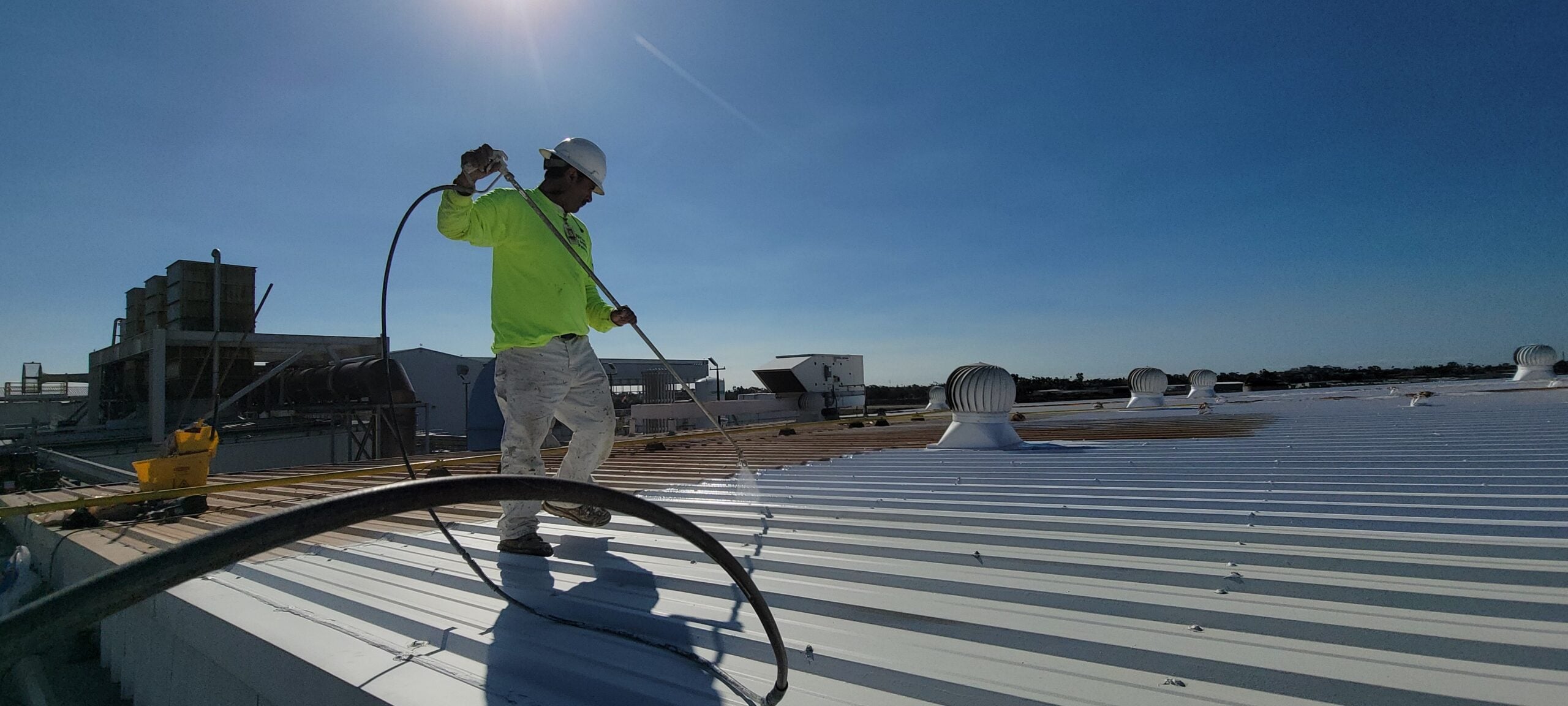 A worker in a high-visibility yellow shirt and hard hat uses a sprayer to coat a large, white industrial metal roof.