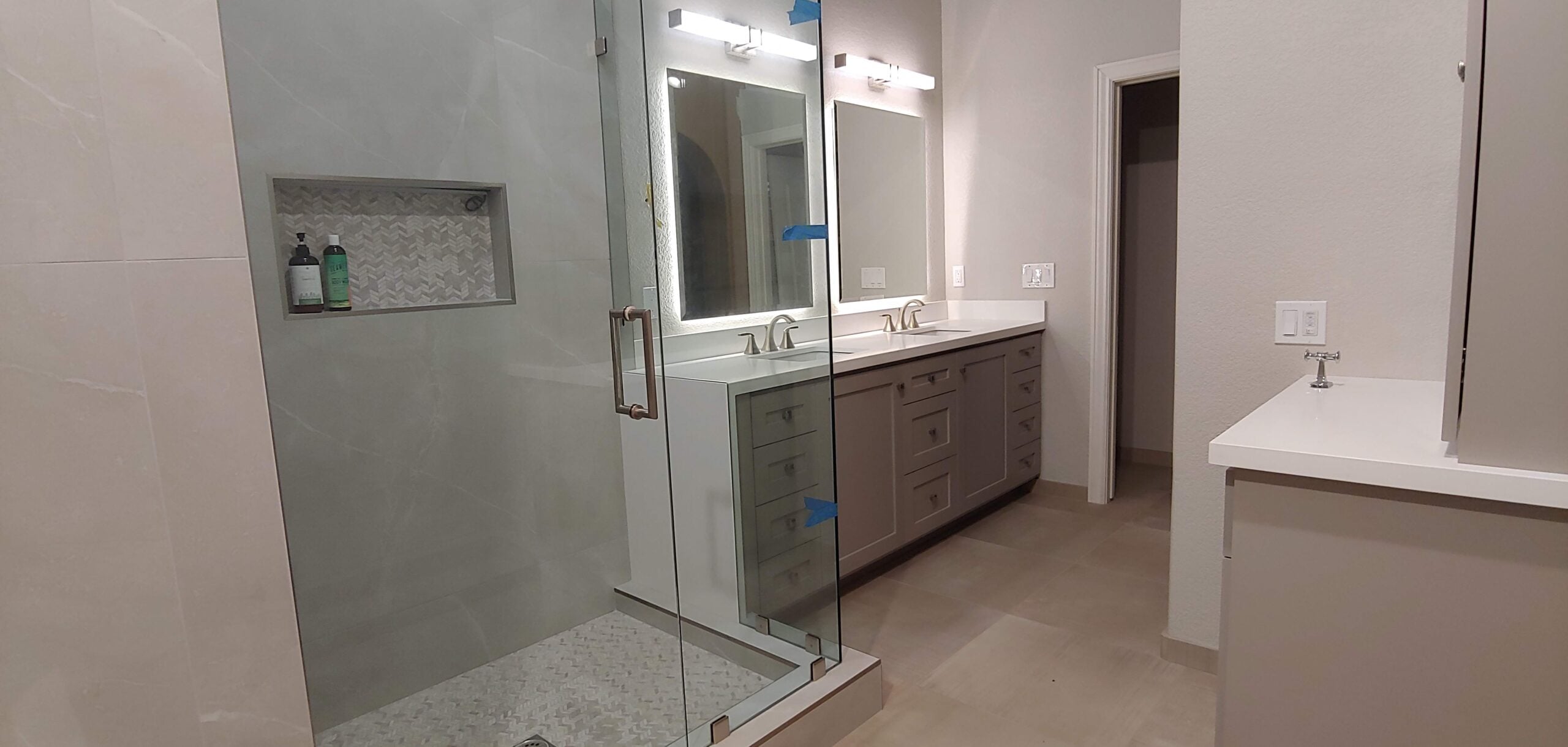 A modern bathroom featuring a walk-in glass shower with a tiled niche, a double vanity with grey cabinets and white countertops, and backlit mirrors.