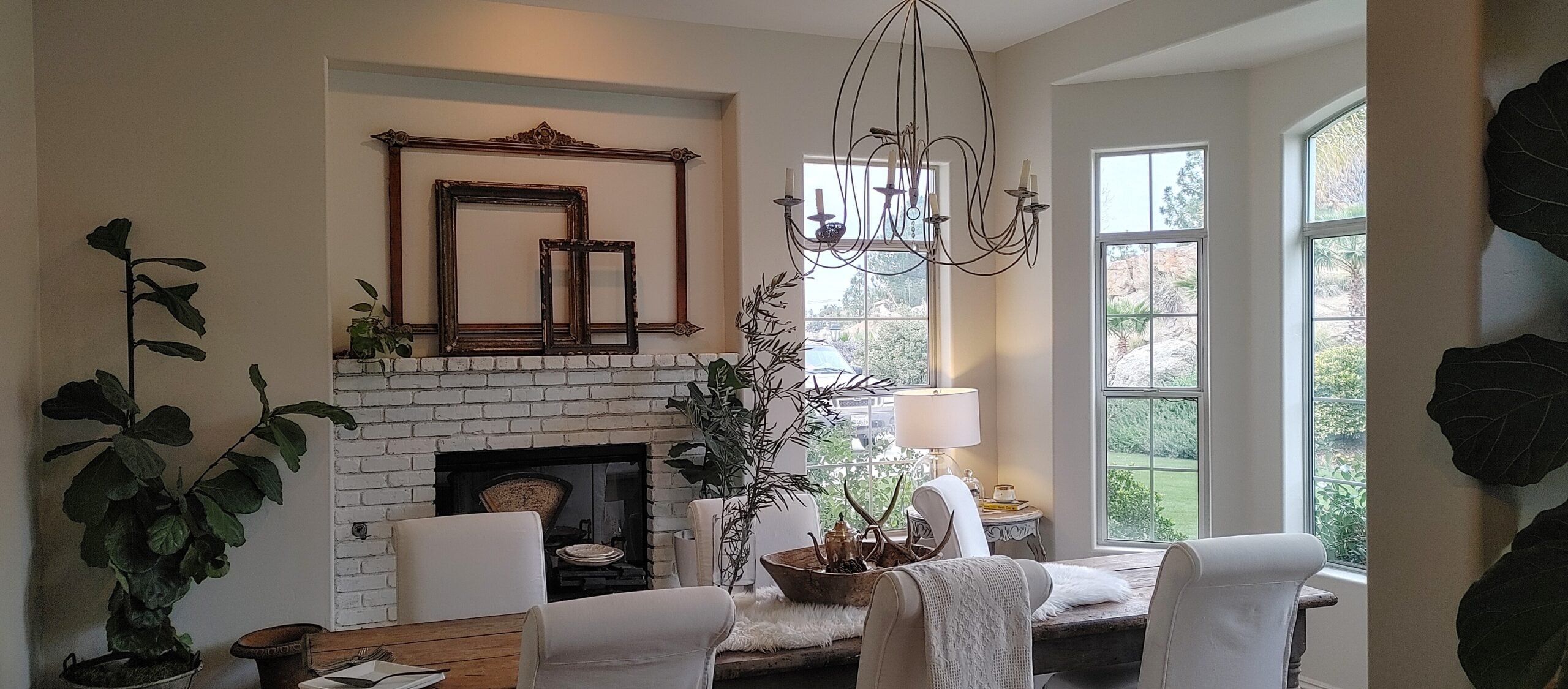 An elegant dining room interior featuring a rustic wooden dining table surrounded by white upholstered chairs. In the background, a white painted brick fireplace is decorated with empty vintage wooden frames stacked inside a wall recess. A unique, thin metal wire chandelier hangs overhead, a tall fiddle-leaf fig plant sits in the corner, and a bay window on the right fills the room with natural light.