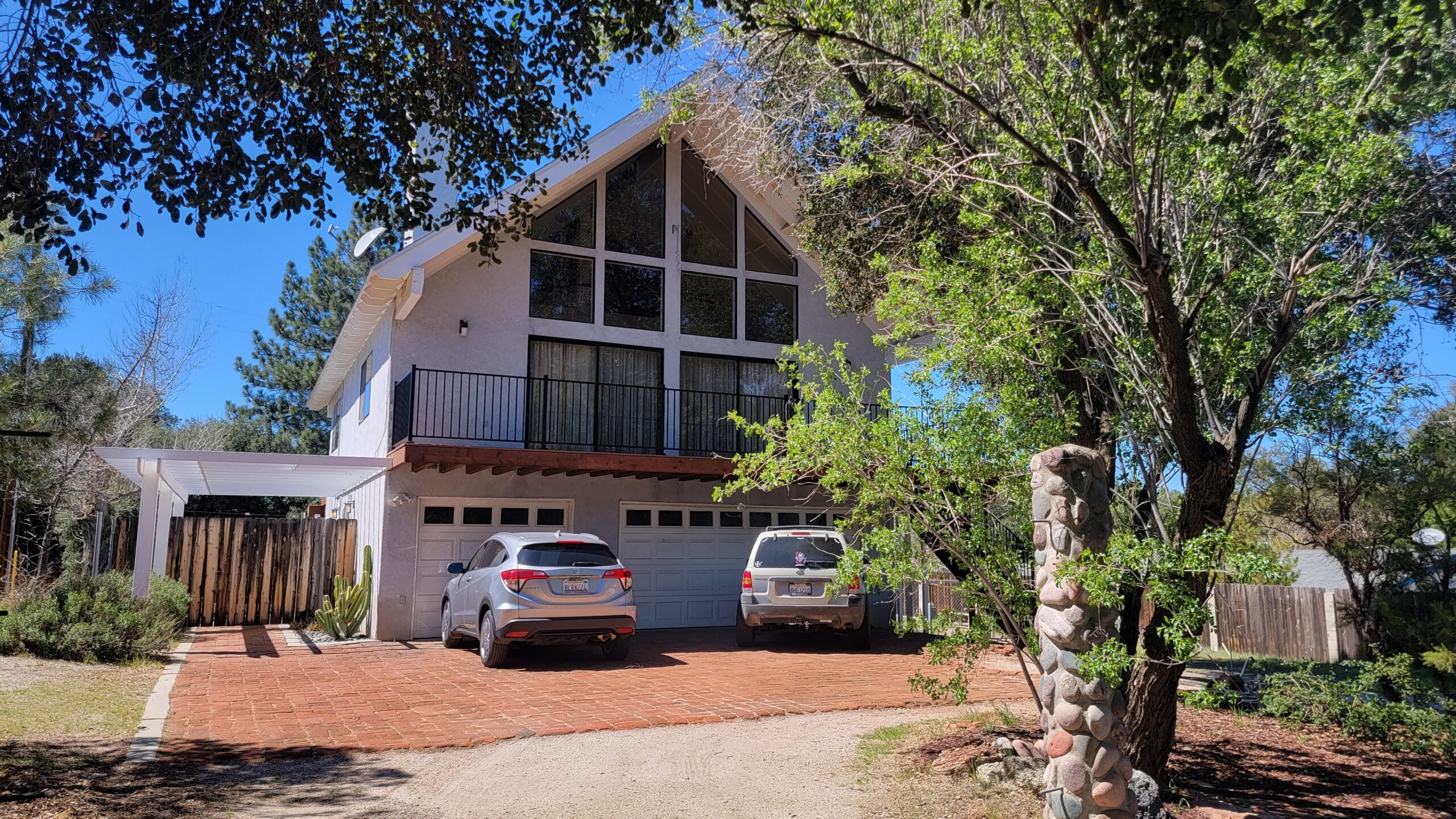 Exterior view of a modern white A-frame house with a brick driveway, parked cars, and large upper-level windows.