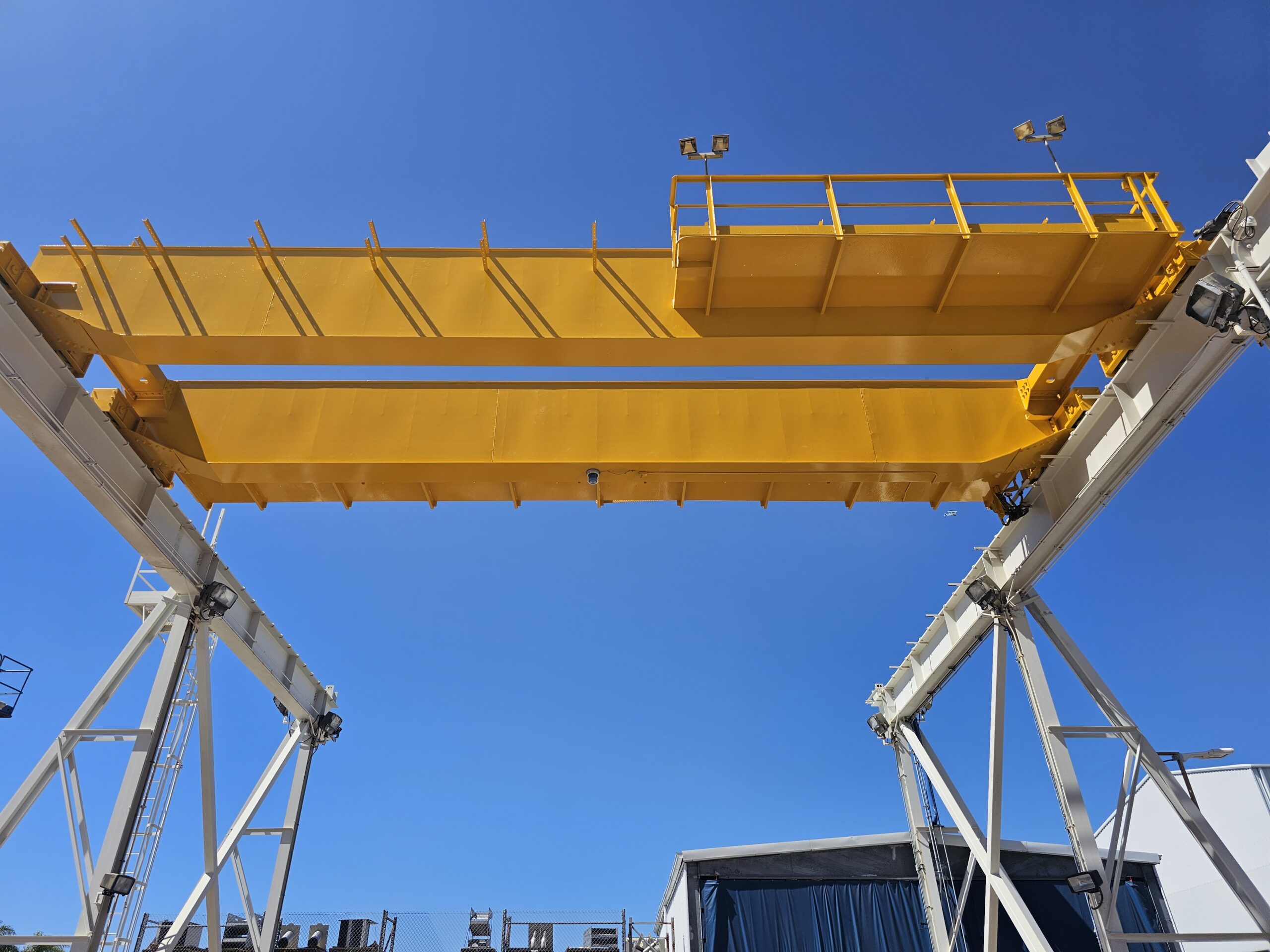 An outdoor industrial overhead crane with large yellow beams against a blue sky.