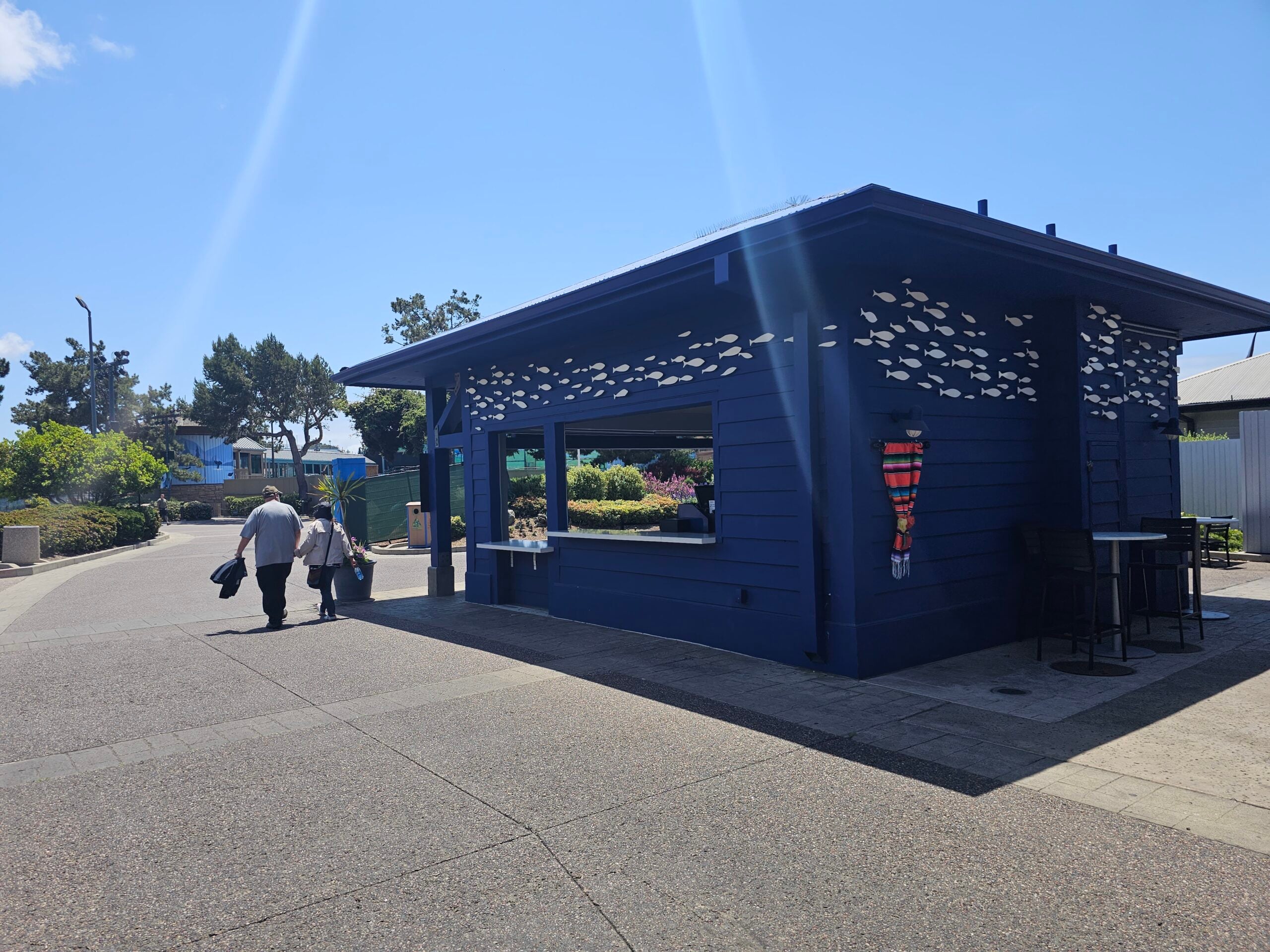 A dark blue outdoor concession booth decorated with white fish patterns, with people walking by on a sunny day.