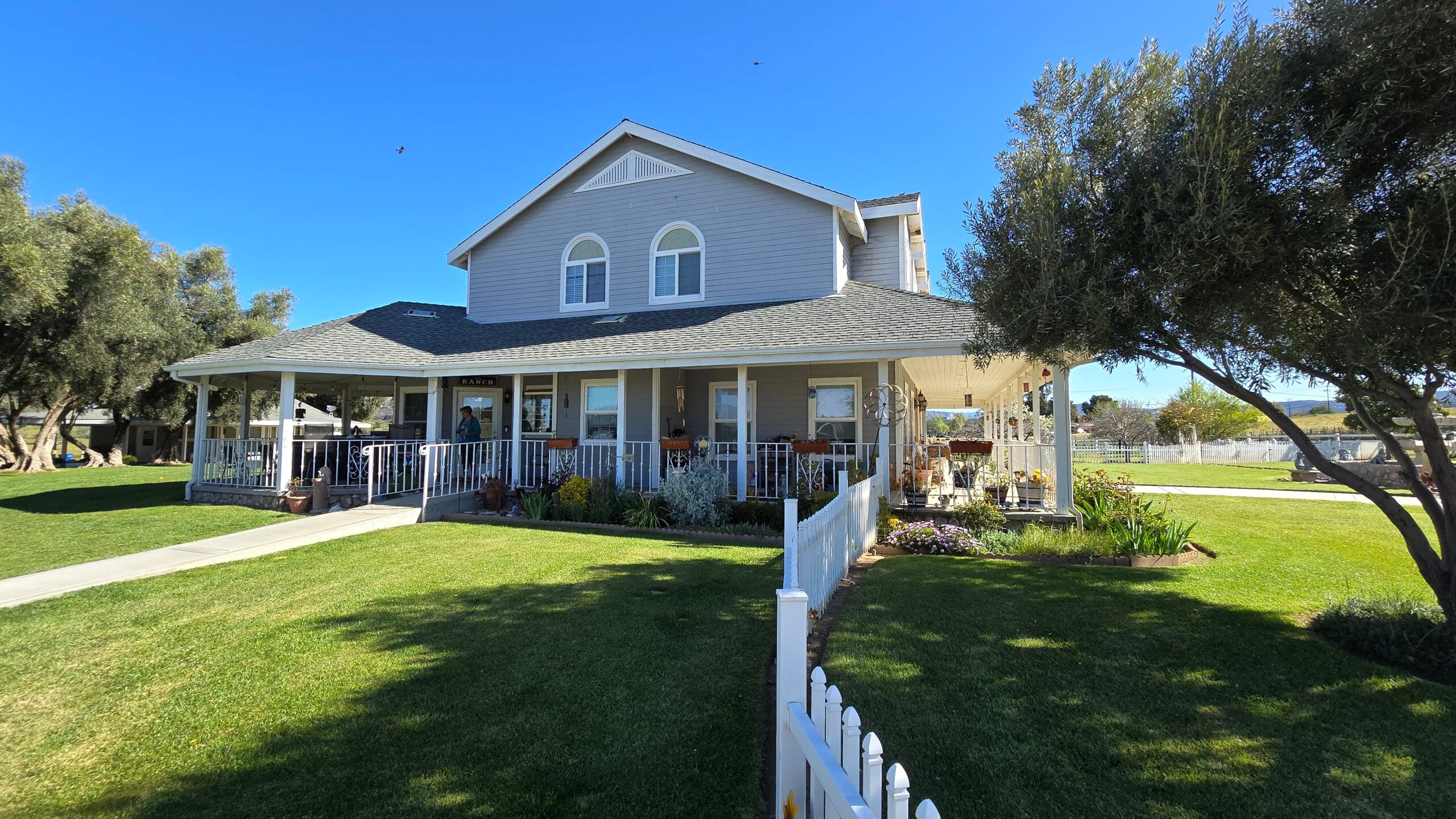 A grey ranch-style house with a spacious front porch and a white picket fence on a sunny day.