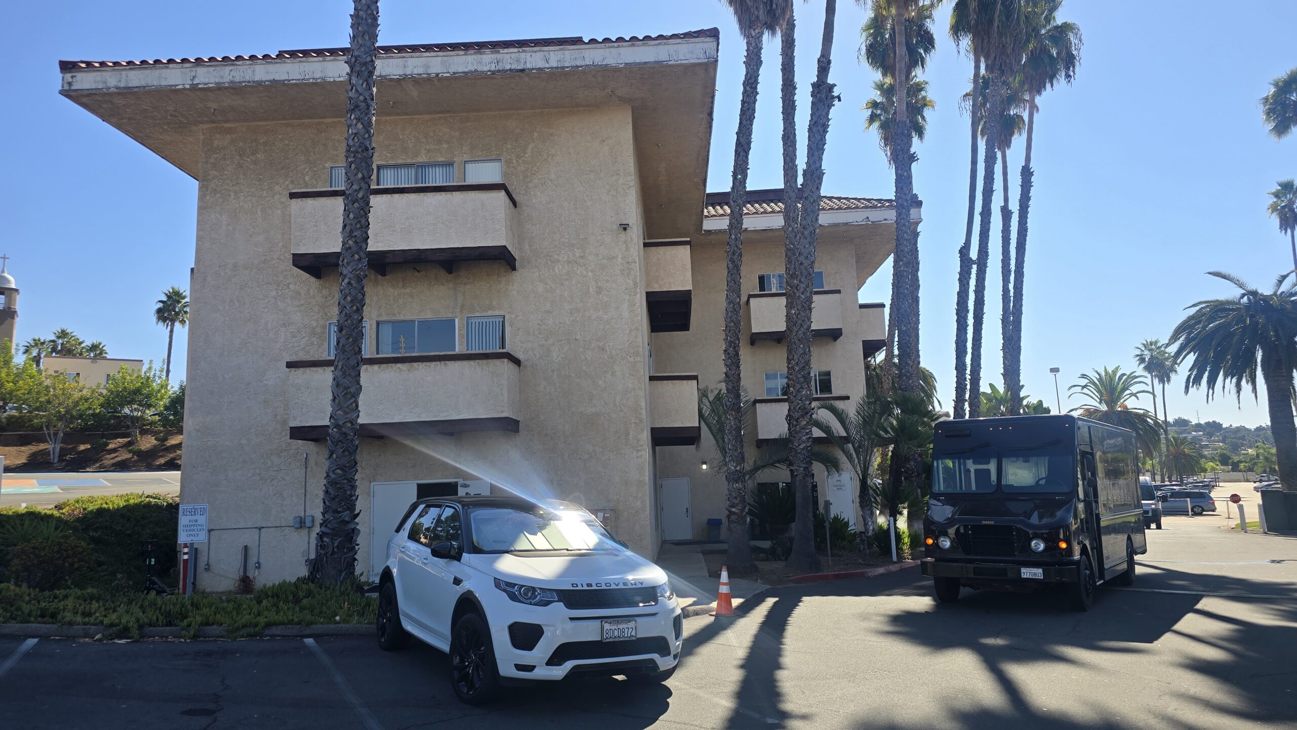 Exterior view of a beige building with palm trees, a white SUV, and a black delivery truck.