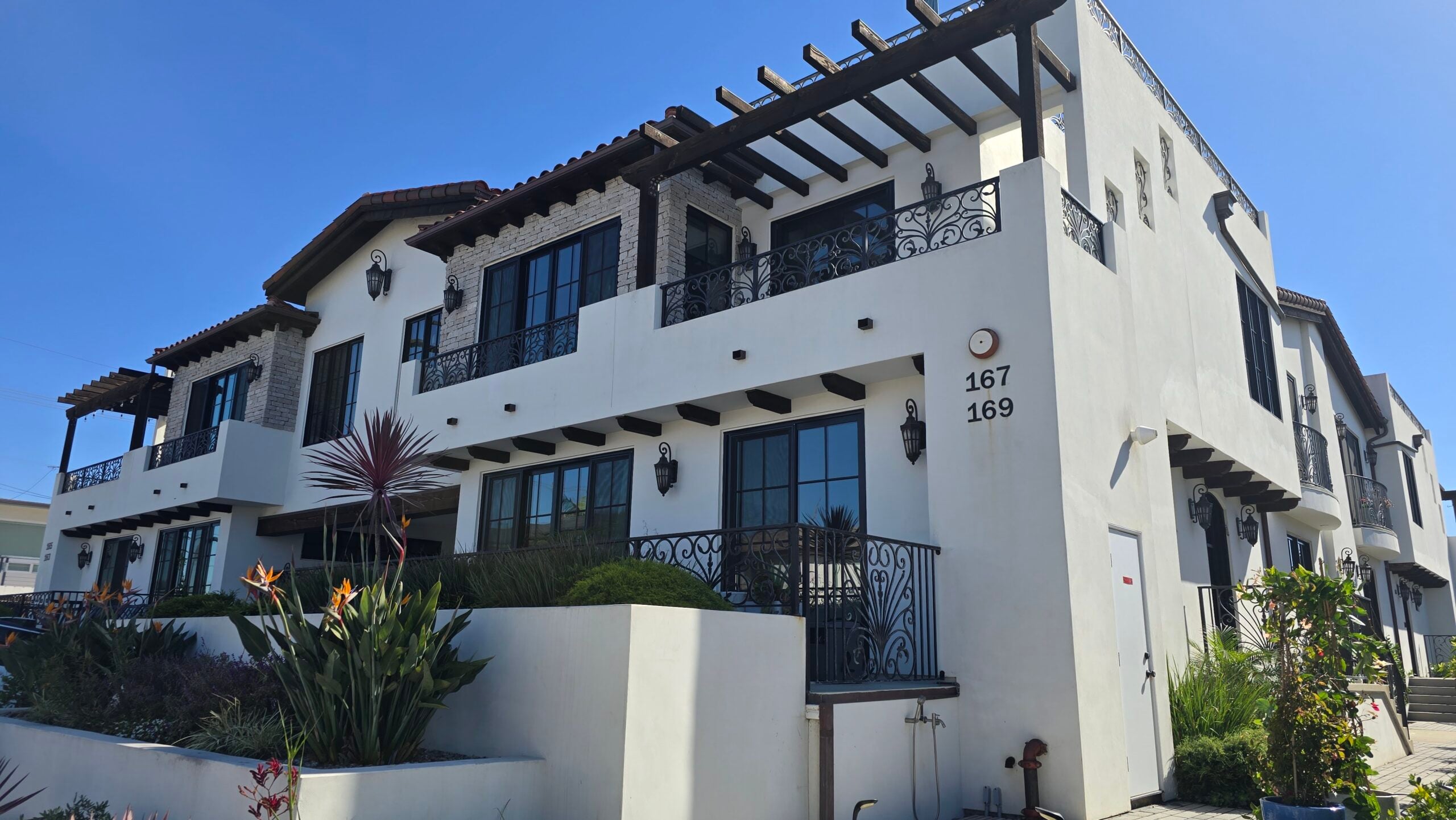 Exterior view of a modern white multi-family residential building with decorative black balconies, wooden pergolas, and front landscaping.