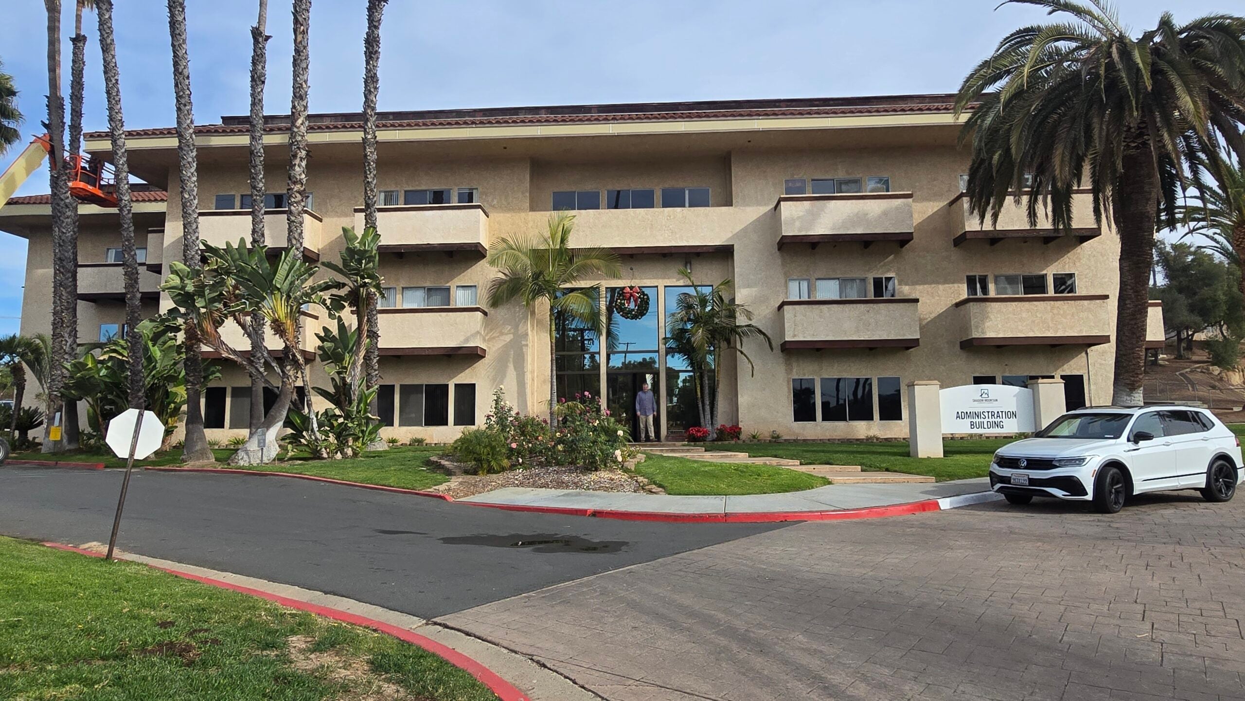 Exterior view of an administration building with palm trees and a parked white car under a cloudy sky.