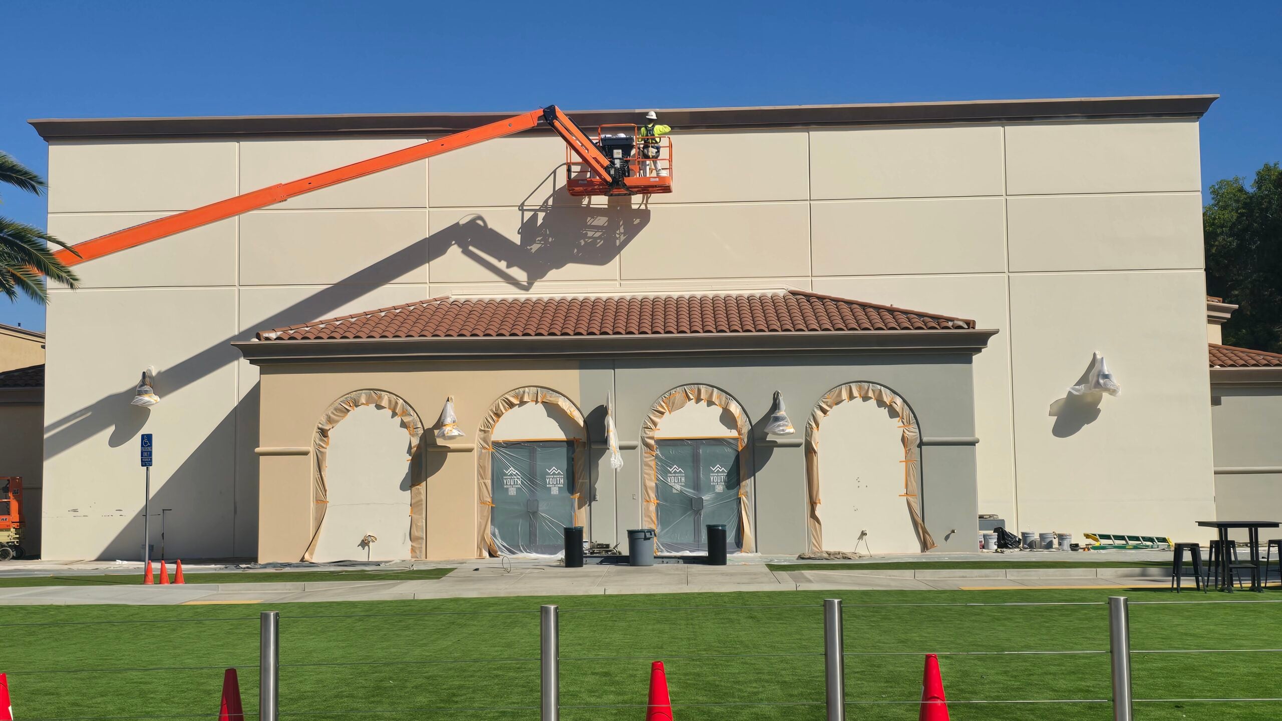 A construction worker using an orange boom lift to paint a large building facade under a blue sky.