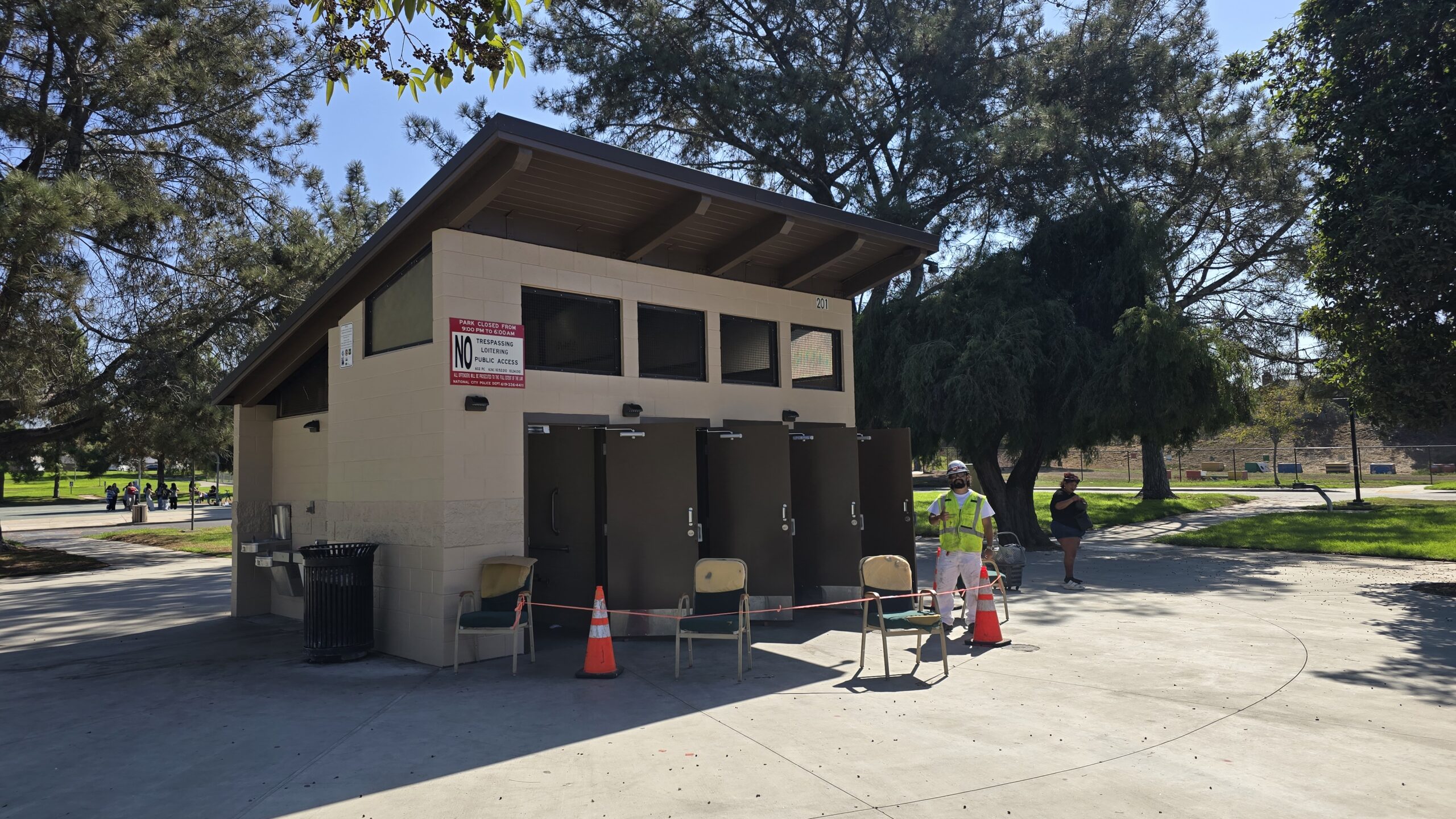 Exterior view of a public park restroom facility undergoing maintenance with a worker and safety cones out front.