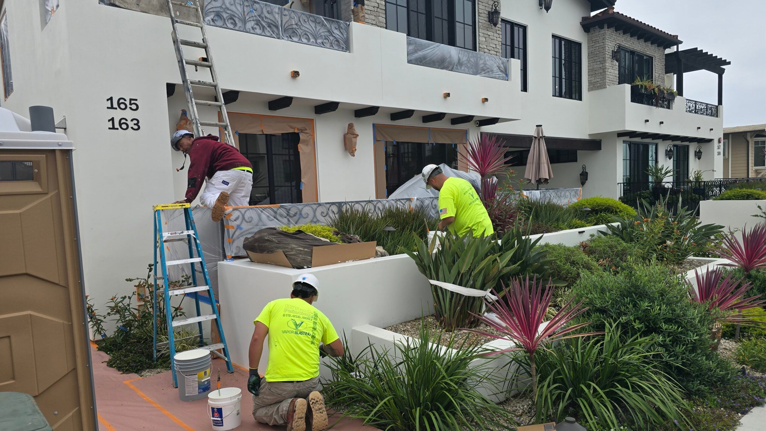 A team of painters working on the front exterior facade and landscaping area of a residential building.
