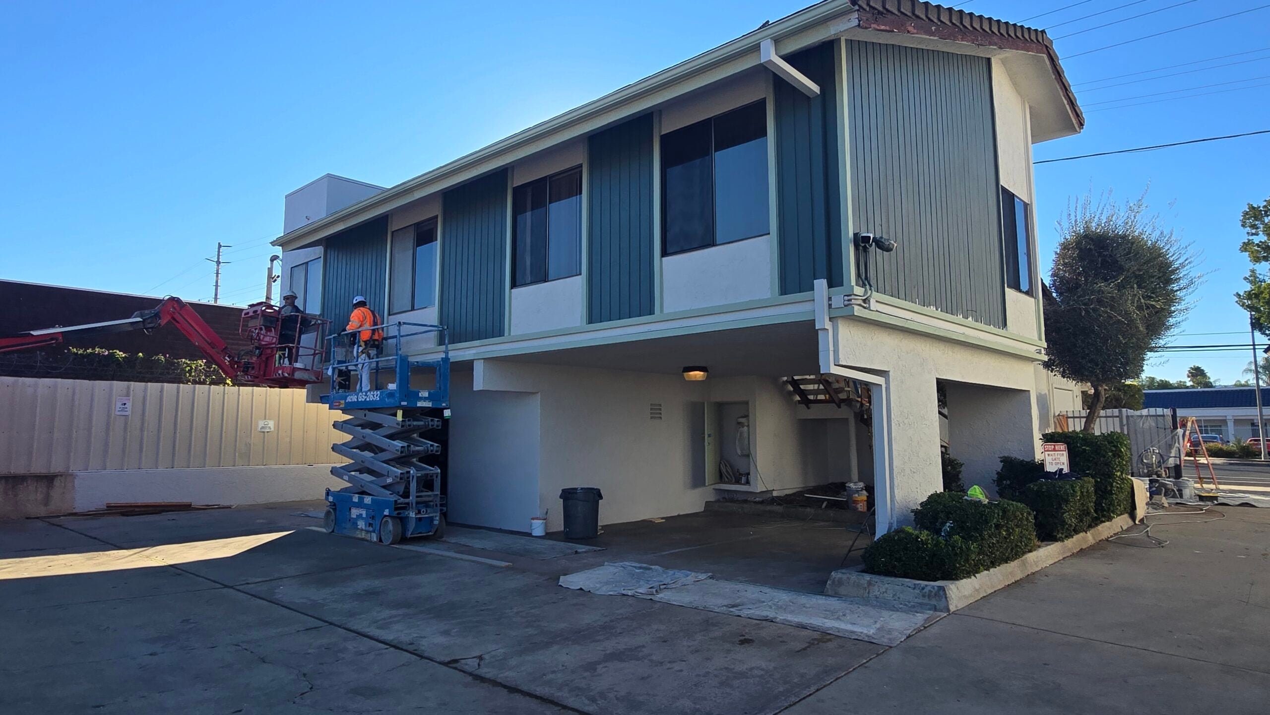 A two-story building with blue-green siding being painted by workers on a blue scissor lift.