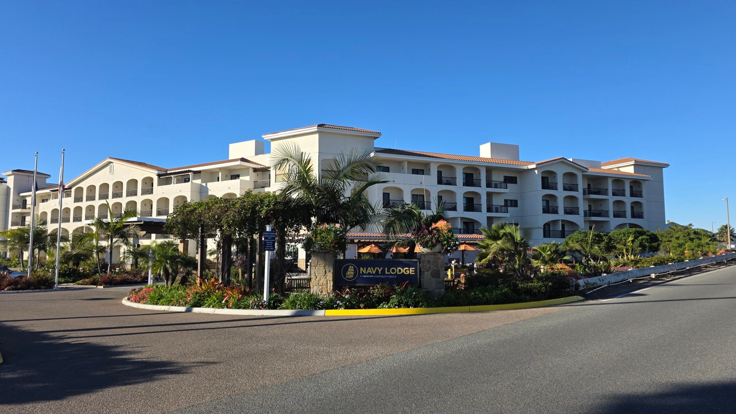 A wide exterior shot of the Navy Lodge hotel building under a clear blue sky. The multi-story resort-style building features a white facade with numerous arched balconies and a red tile roof. In the foreground, a landscaped island with green bushes, palm trees, and flowers holds a dark sign with gold lettering that reads "NAVY LODGE". An asphalt road wraps around the front of the property, and two tall flagpoles stand on the far left side.