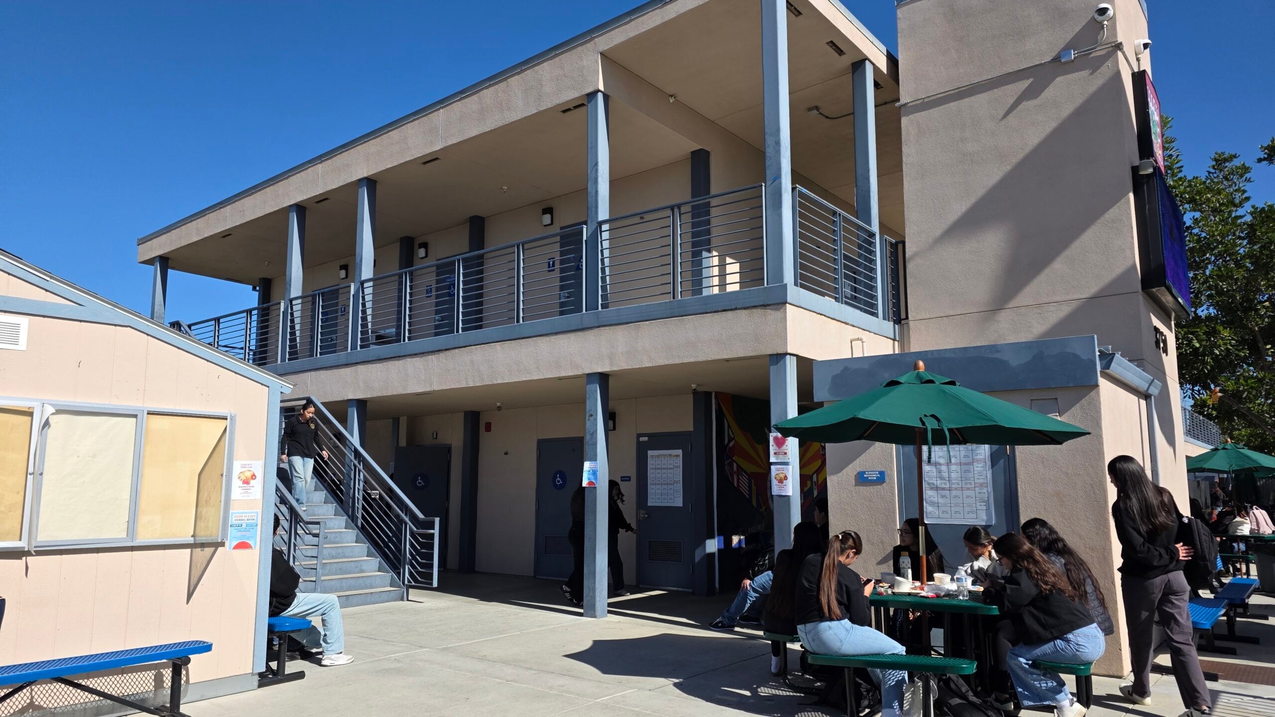 Exterior view of a multi-story school campus building with students sitting at outdoor picnic tables.