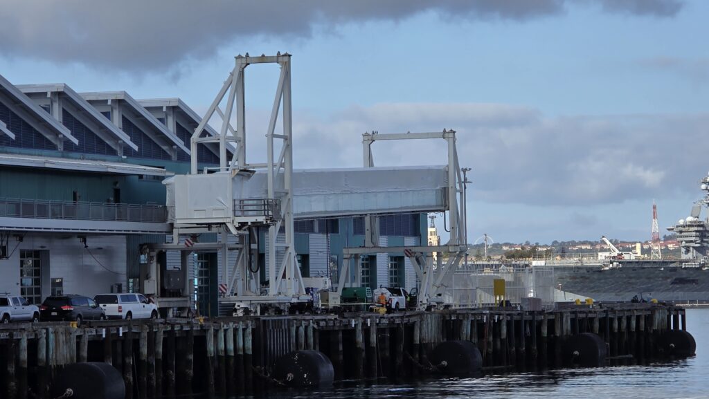 An eye-level shot of a port facility or cruise ship terminal building situated along a wooden pier on a cloudy day. Large white mechanical passenger boarding bridges (jetways) stand ready on the dock. Several vehicles, including a white van, a dark SUV, and a pickup truck, are parked next to the blue-grey building on the left. In the background across the water, a naval ship or large vessel is partially visible on the right under an overcast sky.