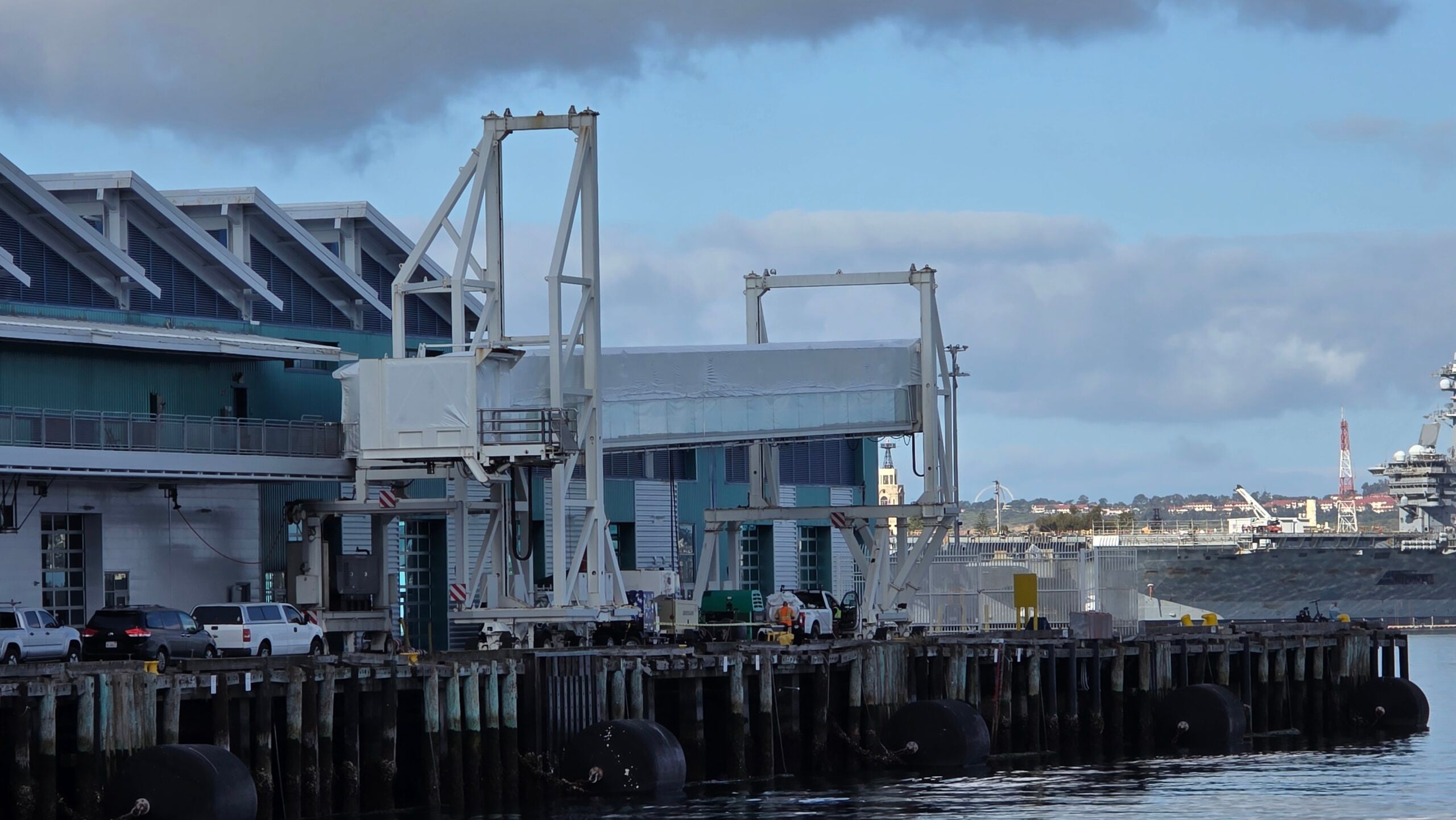 An eye-level shot of a port facility or cruise ship terminal building situated along a wooden pier on a cloudy day. Large white mechanical passenger boarding bridges (jetways) stand ready on the dock. Several vehicles, including a white van, a dark SUV, and a pickup truck, are parked next to the blue-grey building on the left. In the background across the water, a naval ship or large vessel is partially visible on the right under an overcast sky.