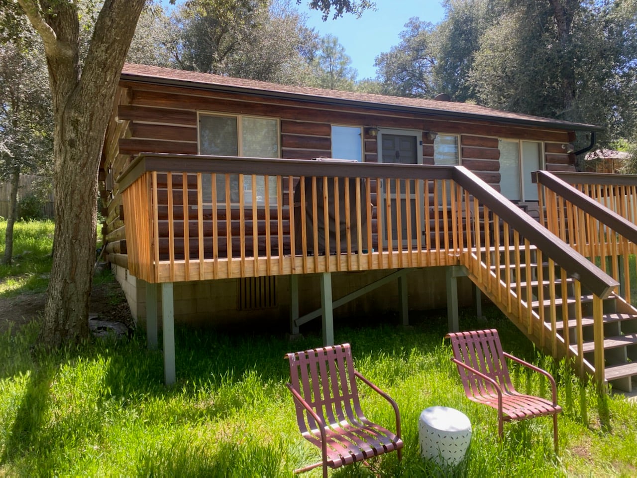 The exterior view of a rustic brown log cabin with an elevated wooden deck and stairs. In the foreground, two reddish-brown metal lawn chairs and a small white side table sit on a sunny green grass lawn, surrounded by mature trees under a clear sky.