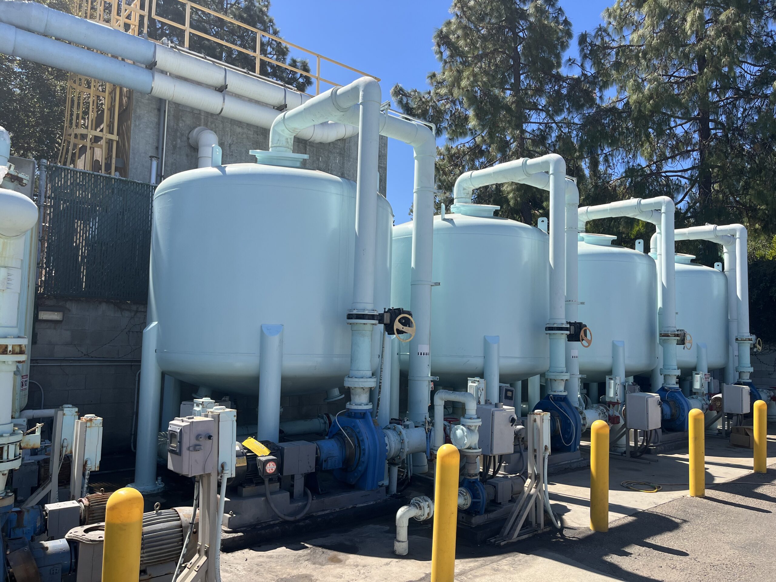 A series of large, light blue industrial filtration tanks connected by white piping, positioned in an outdoor facility with yellow safety bollards in the foreground.