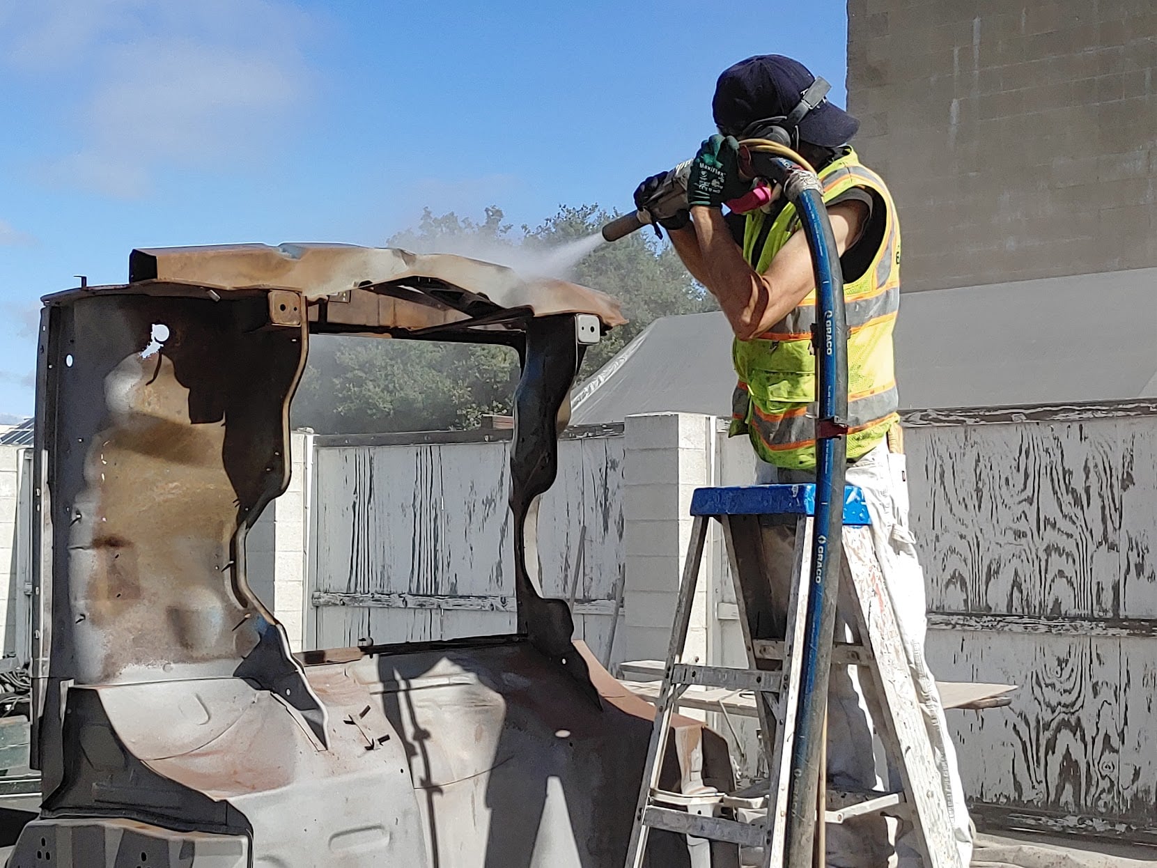 A worker wearing a high-visibility vest and protective gear stands on a ladder while sandblasting a metal vehicle frame.