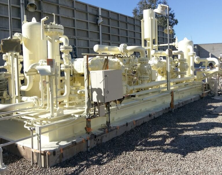 A large, light-colored industrial machine with complex piping and control boxes sits on a concrete pad in an outdoor facility.
