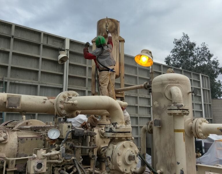 A worker in a hard hat and protective clothing works on an elevated component of industrial machinery at a facility, with pipes and a pressure gauge visible in the foreground.