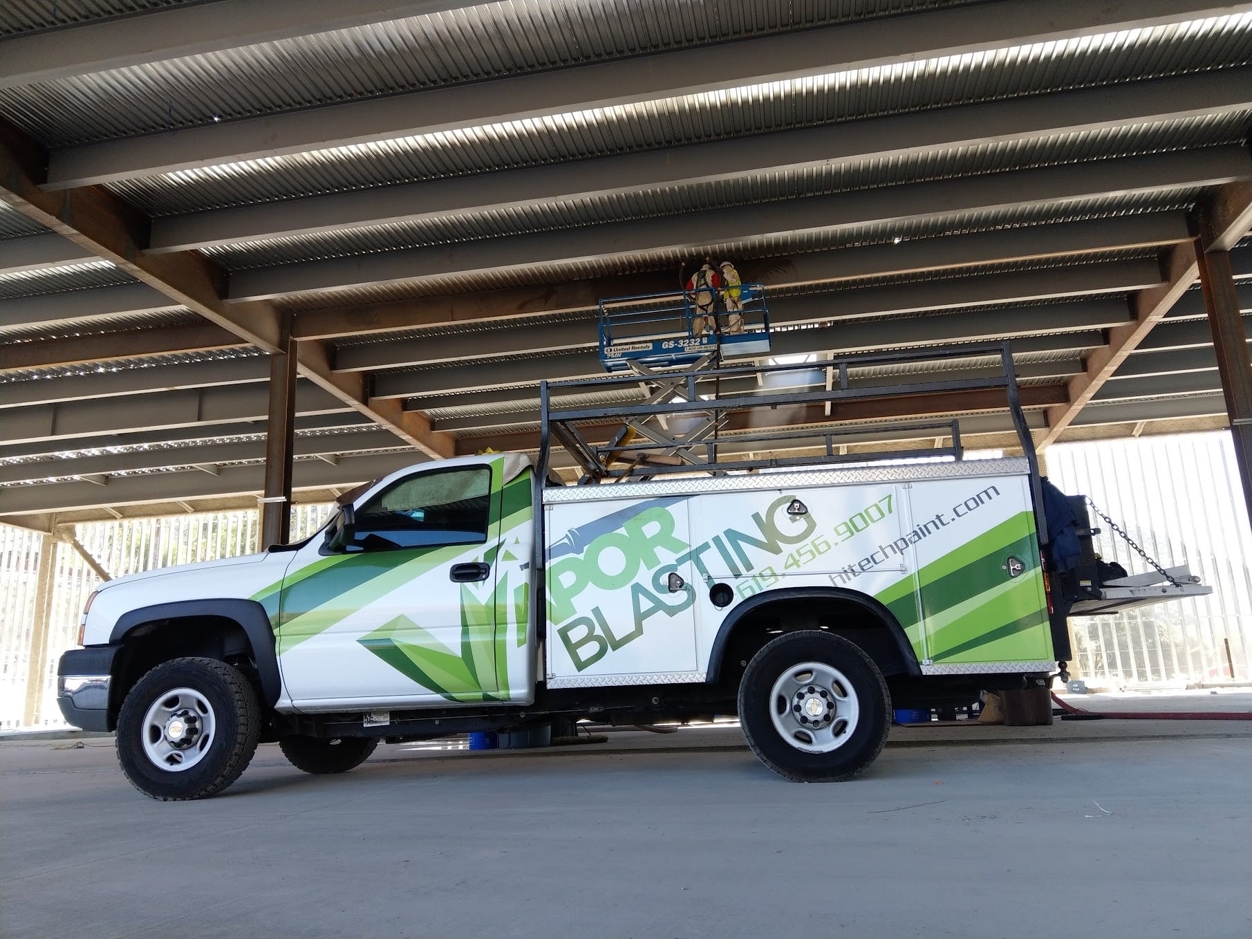 A white utility truck branded with "Vapor Blasting" parked under a large steel structure, where workers are using a scissor lift to work on the ceiling beams.