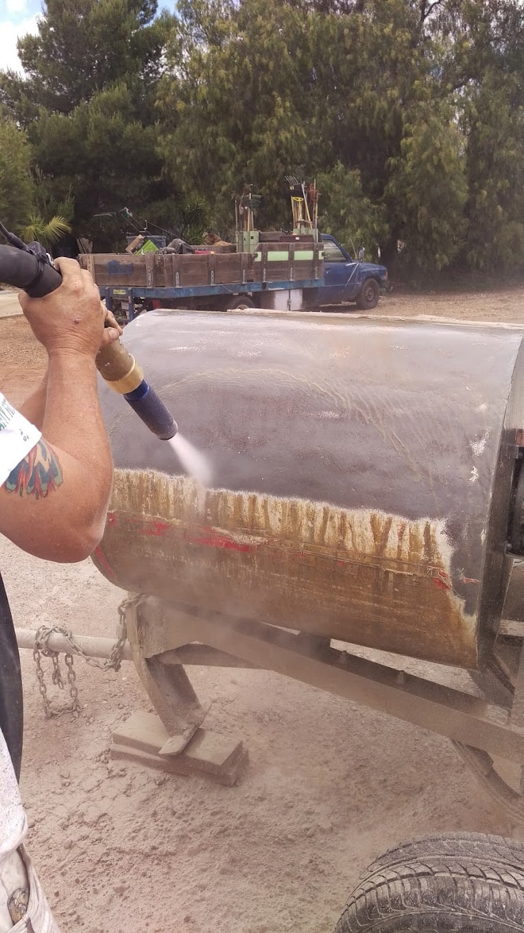 A worker power washing a large cylindrical industrial tank on a trailer, with a flatbed truck parked in the background.
